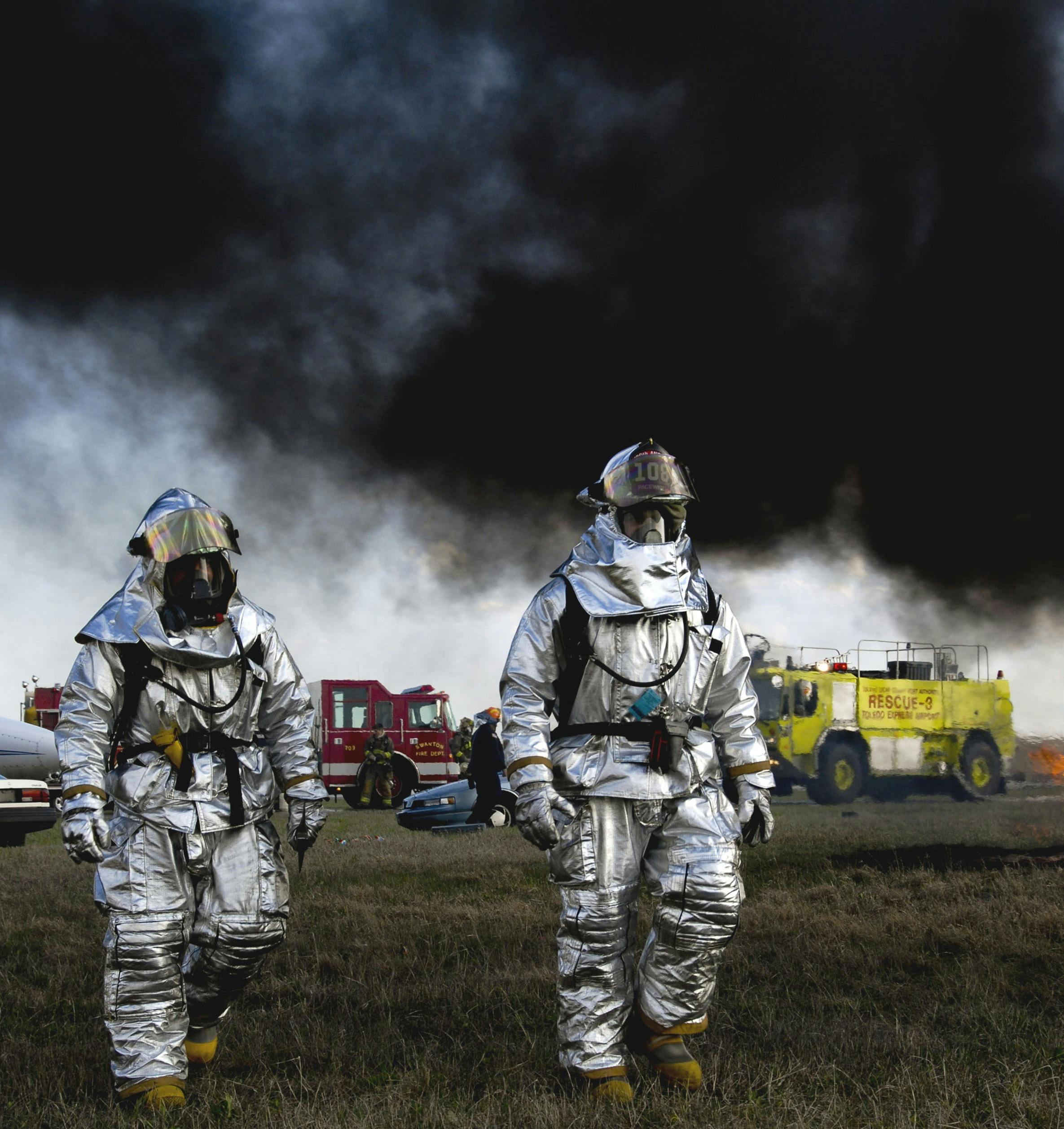 Firefighters in aluminized suits walk amidst smoke and rescue vehicles at a fire scene.